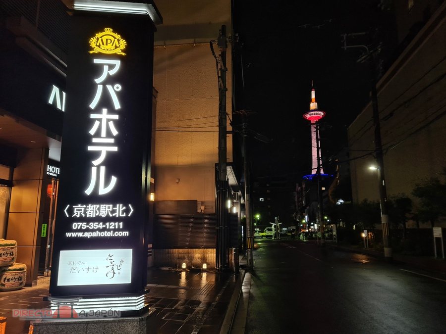 Cartel luminoso de un APA Hotel en Kioto, de noche, con la Torre de Kioto iluminada al fondo.