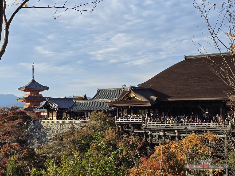 El Hondo de Kiyomizudera con su enorme tejado de corteza de ciprés y una pagoda roja al fondo a la izquierda, entre árboles de otoño.