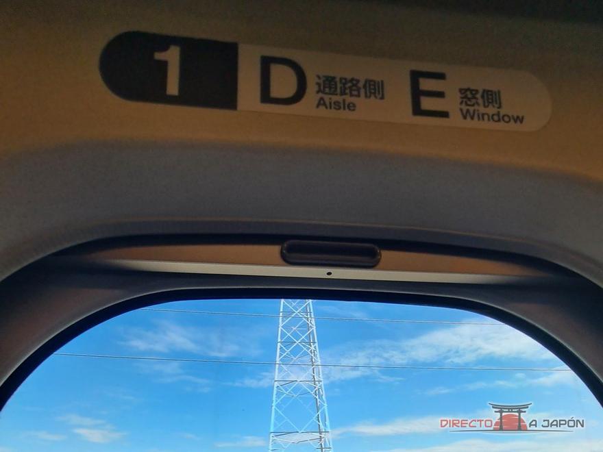 Cartel sobre un asiento del shinkansen con la fila 1 y las letras D (pasillo, Aisle) y E (ventanilla, Window), y el paisaje por la ventanilla.