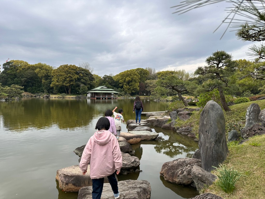 Piedras para cruzar el estanque del jardín Kiyosumi, con rocas, pinos y un pabellón junto al agua al fondo.
