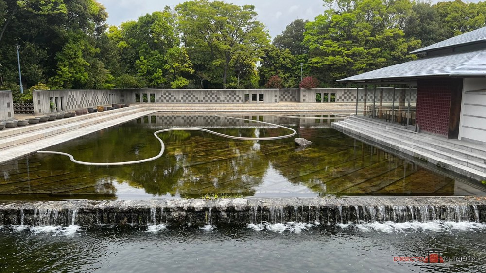 Estanque rectangular con líneas curvas, cascada baja y edificio en Shirotori