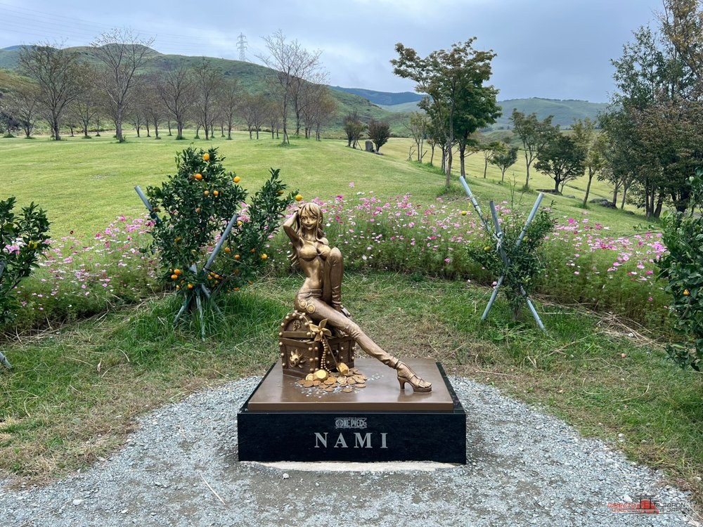 Estatua de Nami sobre un pedestal, rodeada de césped y flores, con las montañas al fondo