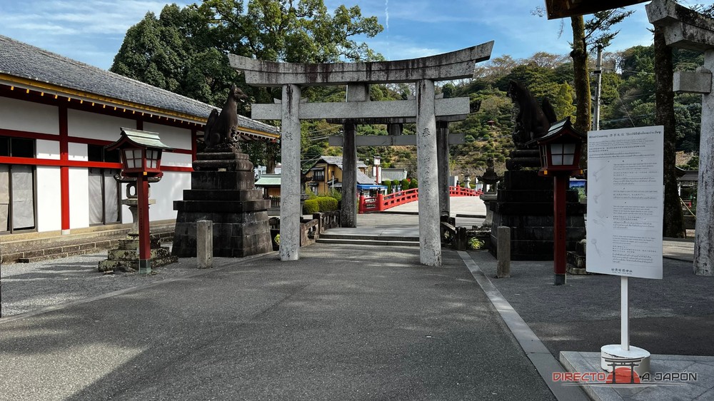 Torii de piedra con zorros de piedra a los lados y un puente rojo al fondo en la entrada del Yutoku Jinja