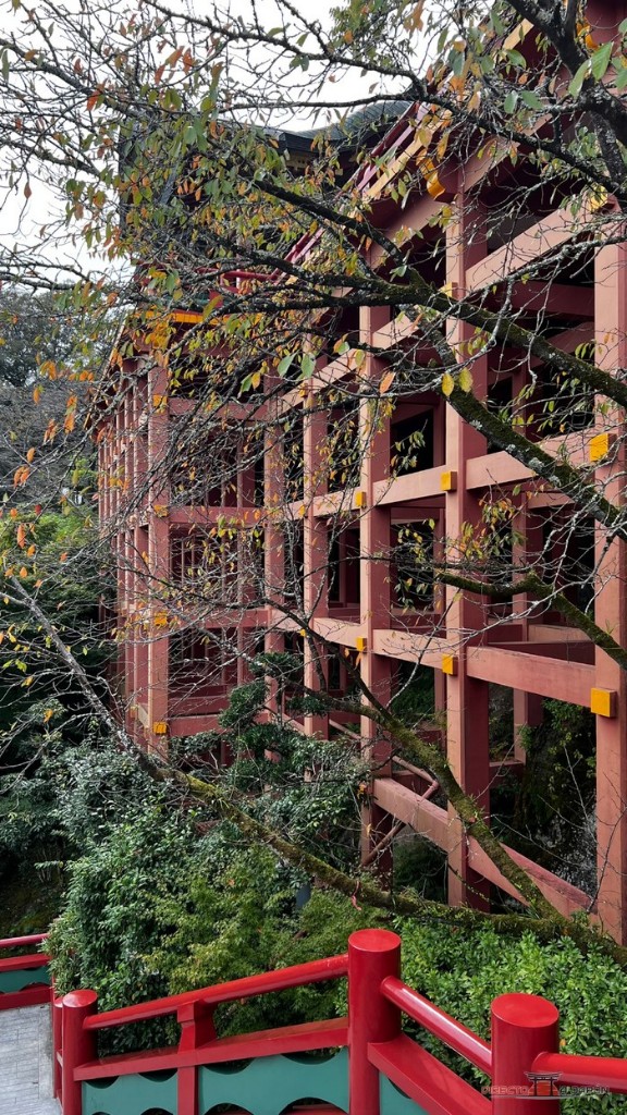 Estructura de vigas y pilares de madera roja del edificio principal del santuario vista desde un lado, entre las ramas de un árbol