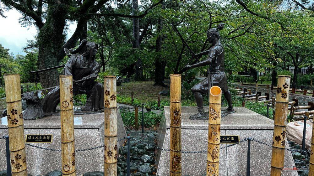 Estatuas de bronce de dos espadachines con las manos en la empuñadura, detrás de unos postes de bambú, junto a la entrada del castillo de Kokura