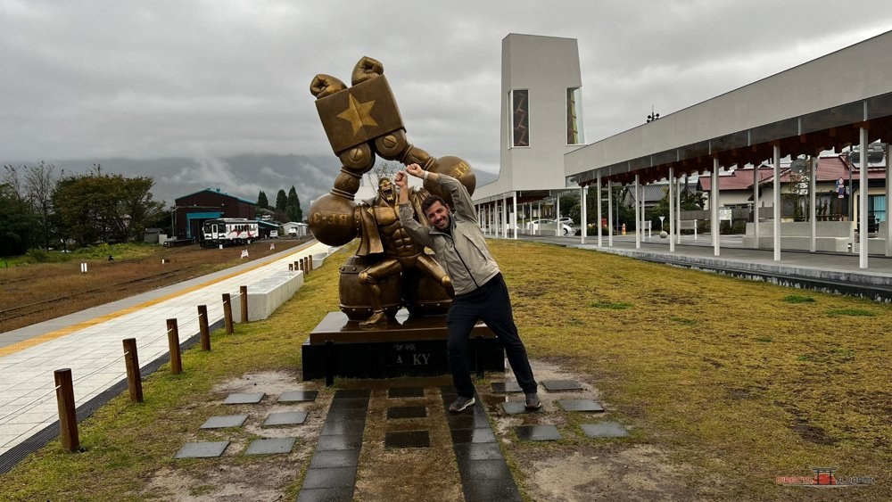 Estatua de Franky junto a una persona en la estación de Takamori, con los raíles de juguete en primer plano